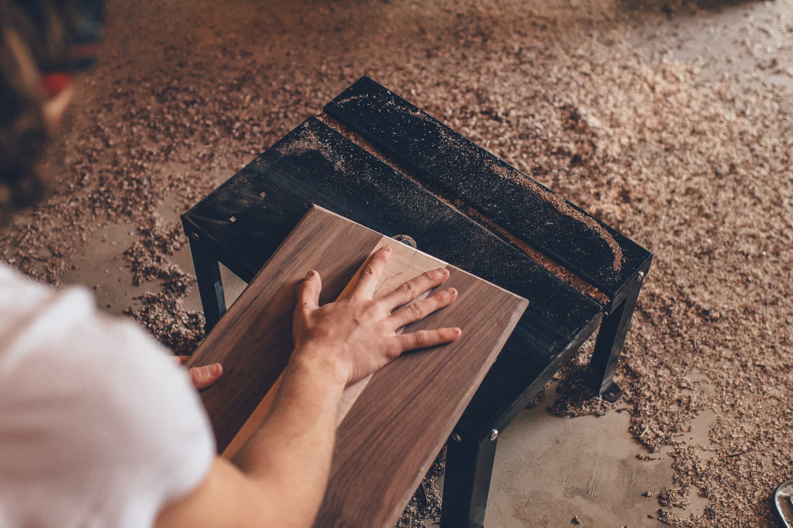 Tangan menekan papan kayu walnut di meja kerja workshop, lantai tertutup serbuk gergaji